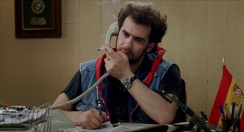 Movie still from “Kika” (1993), directed by Pedro Almodóvar – A man sitting at a table smoking a cigarette and talking on a phone; Medium shot, High angle