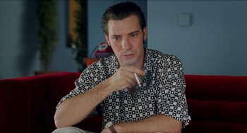 Movie still from “Kika” (1993), directed by Pedro Almodóvar – A man sitting on top of a red couch with a cigarette in his mouth; Close Up shot, Low angle