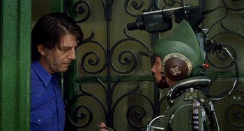 Movie still from “Kika” (1993), directed by Pedro Almodóvar – A man and a woman are standing next to each other in front of an iron gate; Medium shot, Over the shoulder angle