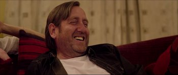 Movie still from “Kill List” (2011), directed by Ben Wheatley – A person sitting on a couch smiling; Close Up shot, Over the shoulder angle