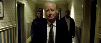 Movie still from “Kill List” (2011), directed by Ben Wheatley – A man in a suit and tie standing in a hallway; Medium shot, Low angle