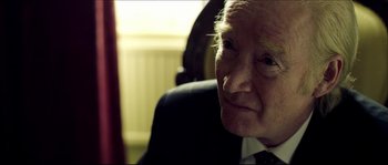Movie still from “Kill List” (2011), directed by Ben Wheatley – An older man wearing a suit sitting in a chair; Close Up shot, Over the shoulder angle