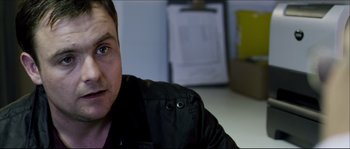 Movie still from “Kill List” (2011), directed by Ben Wheatley – A man sitting on a table with a black jacket on; Close Up shot, Over the shoulder angle