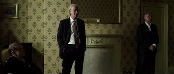 Movie still from “Kill List” (2011), directed by Ben Wheatley – A man in a suit standing in front of a fire place; Medium shot, Low angle