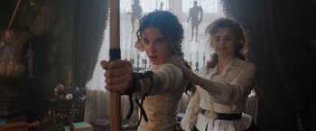Movie still from “Enola Holmes” (2020), directed by Harry Bradbeer – A young woman holding a bow and arrow in front of another young woman; Medium shot, Over the shoulder angle
