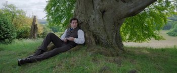 Movie still from “Enola Holmes” (2020), directed by Harry Bradbeer – A man sitting on the ground next to a tree; Medium shot, High angle