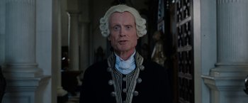 Movie still from “Enola Holmes” (2020), directed by Harry Bradbeer – A man in a wig and wig stands in front of a statue; Close Up shot, Low angle
