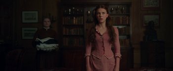 Movie still from “Enola Holmes” (2020), directed by Harry Bradbeer – A woman in a pink dress standing in front of bookshelves; Medium shot, Over the shoulder angle