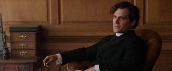 Movie still from “Enola Holmes” (2020), directed by Harry Bradbeer – A man sitting in a chair wearing a suit and tie; Close Up shot, Low angle