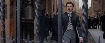 Movie still from “Enola Holmes” (2020), directed by Harry Bradbeer – A man in a suit and tie standing in front of a fence; Medium shot, Low angle