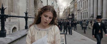 Movie still from “Enola Holmes” (2020), directed by Harry Bradbeer – A woman is reading a paper while standing on the street; Medium shot, Over the shoulder angle