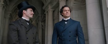 Movie still from “Enola Holmes” (2020), directed by Harry Bradbeer – A man in a suit and a hat standing next to another man in a suit; Medium shot, Over the shoulder angle