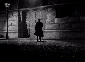Movie still from “Kind Hearts and Coronets” (1949), directed by Robert Hamer – A man standing in front of a wall with a bag; Wide shot, Low angle