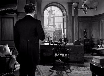Movie still from “Kind Hearts and Coronets” (1949), directed by Robert Hamer – A man standing in front of a desk in front of a window; Wide shot, Over the shoulder angle