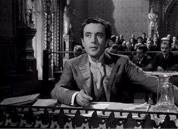 Movie still from “Kind Hearts and Coronets” (1949), directed by Robert Hamer – A black and white photo of a man sitting at a table in a courtroom; Medium shot, Low angle