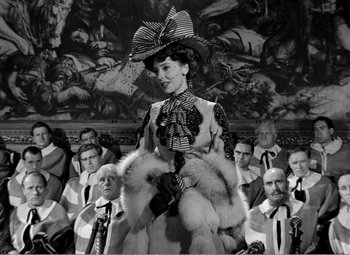 Movie still from “Kind Hearts and Coronets” (1949), directed by Robert Hamer – A woman in a hat and a fur coat standing in front of a group of people; Medium shot, Over the shoulder angle