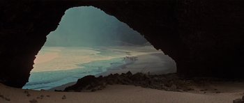 Movie still from “King Kong” (1976), directed by John Guillermin – People are walking on the beach under a large cave; Extreme Wide shot, High angle