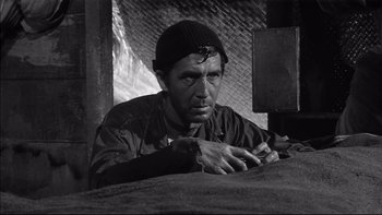 Movie still from “King Rat” (1965), directed by Bryan Forbes – A man sitting on top of a bed in front of a brick wall; Medium shot, Low angle