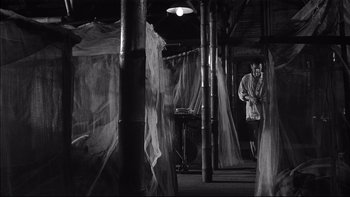 Movie still from “King Rat” (1965), directed by Bryan Forbes – A man standing in front of a lamp in a room; Wide shot, High angle