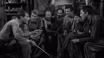 Movie still from “King Rat” (1965), directed by Bryan Forbes – A black and white photo of a group of men sitting in a circle; Medium shot, High angle
