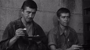 Movie still from “King Rat” (1965), directed by Bryan Forbes – A black and white photo of two men sitting next to each other; Medium shot, High angle