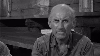 Movie still from “King Rat” (1965), directed by Bryan Forbes – An older man is looking at the camera while sitting in front of a bench; Close Up shot, Low angle