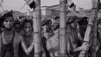 Movie still from “King Rat” (1965), directed by Bryan Forbes – A black and white photo of a group of men standing next to each other in front of a barbed wire fence; Wide shot, High angle