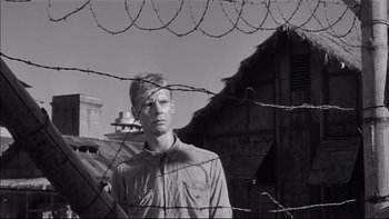 Movie still from “King Rat” (1965), directed by Bryan Forbes – A man standing behind a barbed wire fence; Close Up shot, Low angle