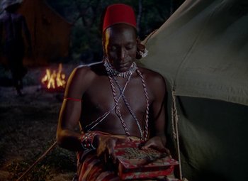 Movie still from “King Solomon's Mines” (1950), directed by Andrew Marton – A man sitting in front of a tent holding a box; Medium shot, High angle