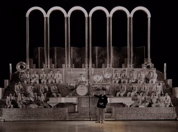 Movie still from “King of Jazz” (1930), directed by Walter Lantz – A person standing in front of a large picture of a crowd; Extreme Wide shot, High angle