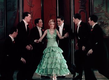 Movie still from “King of Jazz” (1930), directed by Walter Lantz – A woman in a green dress is surrounded by men in black suits; Wide shot, High angle