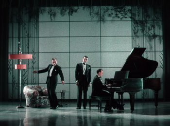 Movie still from “King of Jazz” (1930), directed by Walter Lantz – A group of men in suits and ties standing in front of a piano; Wide shot, Low angle