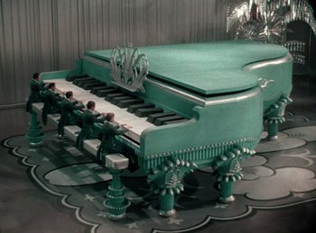 Movie still from “King of Jazz” (1930), directed by Walter Lantz – A green piano sitting on top of a floor next to a wall; Extreme Wide shot, High angle