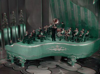 Movie still from “King of Jazz” (1930), directed by Walter Lantz – A group of people sitting on a stage with a man playing a trombone; Extreme Wide shot, High angle
