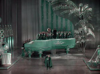 Movie still from “King of Jazz” (1930), directed by Walter Lantz – A group of people standing around a piano; Extreme Wide shot, High angle