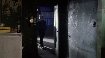Movie still from “King of New York” (1990), directed by Abel Ferrara – A man standing in a dark room in front of an open door; Wide shot, Low angle