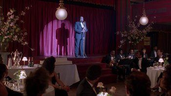 Movie still from “King of New York” (1990), directed by Abel Ferrara – A man standing on a stage in front of a microphone; Wide shot, Over the shoulder angle
