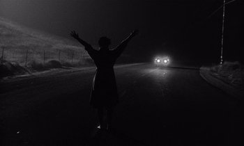 Movie still from “Kiss Me Deadly” (1955), directed by Robert Aldrich – A woman standing on the side of a road with her arms in the air; Wide shot, Low angle