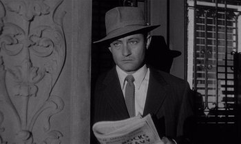 Movie still from “Kiss Me Deadly” (1955), directed by Robert Aldrich – A black and white photo of a man in a suit and hat holding a newspaper; Close Up shot, Low angle