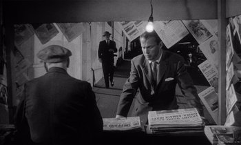 Movie still from “Kiss Me Deadly” (1955), directed by Robert Aldrich – A man in a suit and tie standing next to a pile of newspapers; Medium shot, Over the shoulder angle