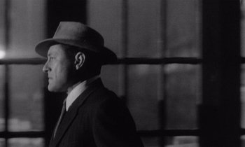 Movie still from “Kiss Me Deadly” (1955), directed by Robert Aldrich – A man wearing a suit and a hat standing in front of a window; Close Up shot, Low angle