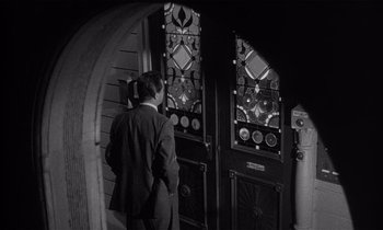 Movie still from “Kiss Me Deadly” (1955), directed by Robert Aldrich – A black and white photo of a man standing in front of a stained glass window; Medium shot, High angle