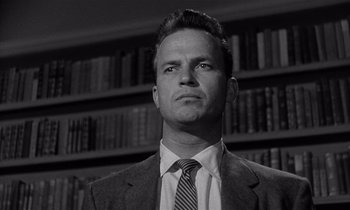 Movie still from “Kiss Me Deadly” (1955), directed by Robert Aldrich – A black and white photo of a man in a suit and tie in front of bookshelves; Close Up shot, Low angle