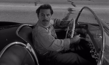 Movie still from “Kiss Me Deadly” (1955), directed by Robert Aldrich – A black and white photo of a man driving a car; Close Up shot, High angle