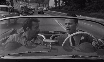 Movie still from “Kiss Me Deadly” (1955), directed by Robert Aldrich – Two men in the back seat of a car on the road; Medium shot, Over the shoulder angle