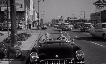 Movie still from “Kiss Me Deadly” (1955), directed by Robert Aldrich – Two people in a car driving down a street; Wide shot, High angle