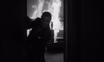 Movie still from “Kiss Me Deadly” (1955), directed by Robert Aldrich – A man standing in front of a fire in a room; Medium shot, Low angle
