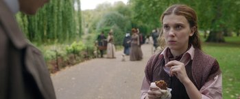 Movie still from “Enola Holmes 2” (2022), directed by Harry Bradbeer – A woman holding a cupcake in her hand; Medium shot, Over the shoulder angle