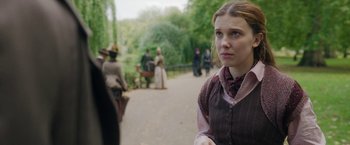 Movie still from “Enola Holmes 2” (2022), directed by Harry Bradbeer – A young woman in a dress standing in a park; Medium shot, Over the shoulder angle