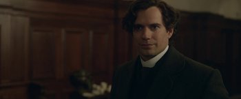 Movie still from “Enola Holmes 2” (2022), directed by Harry Bradbeer – A man in a priest's outfit standing in front of a wooden wall; Close Up shot, Over the shoulder angle